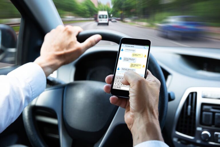 A driver in a white shirt holds a smartphone, texting while steering the wheel