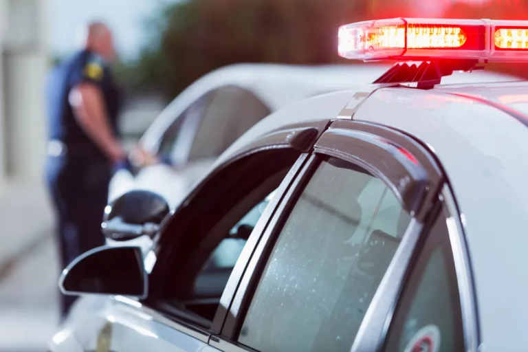 A police vehicle with flashing lights parked beside a car, as an officer approaches the driver’s side